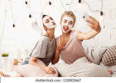 Portrait Of Cute Young Women Sending Kisses Smiling Sweetly And Making Amusing Faces For Picture. Cheerful Lovely Friends Taking Selfie At Home With Facial Cosmetic Mask. Skincare Concept