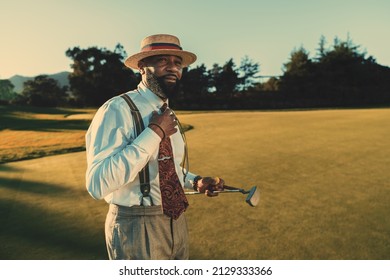 A portrait of a dapper mature black man in an elegant outfit with white a shirt, tie, and trousers with suspenders, standing on an evening golf field and holding his club and a ball; a copy space area