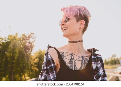 Portrait Of Daring Young Tattooed Woman With Short Pink Hair, Open Chest, Sitting Outdoor In Round Glasses