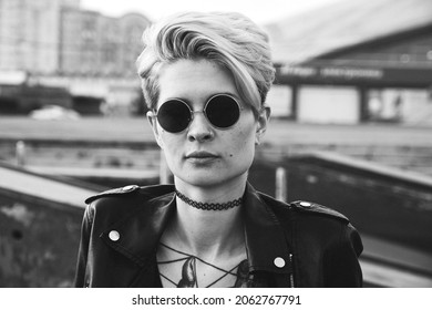 Portrait Of Daring Young Tattooed Woman With Short Hair In Round Glasses Laying On The Fround Outdoor, Looking At The Camera