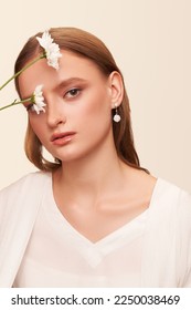 Portrait of a dark blond lady with a gentle makeup. The girl is wearing golden stud earrings made out of pearl beads with pendants made as mother-of-pearl flowers and posing on the light background.  