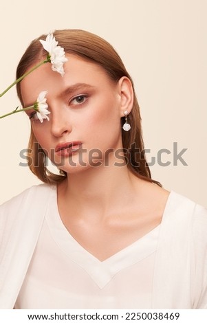 Portrait of a dark blond lady with a gentle makeup. The girl is wearing golden stud earrings made out of pearl beads with pendants made as mother-of-pearl flowers and posing on the light background.  