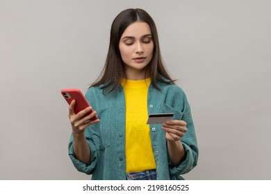Portrait of dark haired concentrated woman holding smart phone and credit card, entering data, online shopping, wearing casual style jacket. Indoor studio shot isolated on gray background. - Shutterstock ID 2198160525