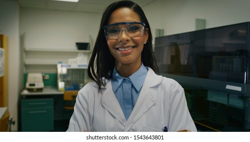 Portrait Of  Dark Skin Female Scientist Wearing Protective Glasses Is Smiling Satisfied With Great Results Of Completed Research In Camera In Laboratory.