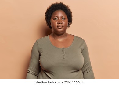 Portrait of dark skinned plump woman with curly hair looks directly at camera has serious expression dressed in casual jumper poses in studio over brown background. Body positive female model indoor - Powered by Shutterstock