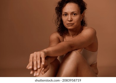Portrait of a dark-haired curly woman with skin flaws, cellulite and stretch marks posing in beige underwear, looking at camera against colored background with copy space. Body positivity concept