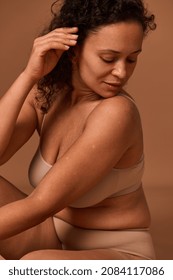 Portrait of a dark-haired curly woman with skin flaws, cellulite and stretch marks posing in beige underwear, looking down against colored background with copy space. Body positive concept