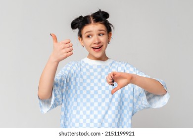 Portrait Of Dark-haired Teen Girl Showing Thumbs Up And Thumbs Down, Like Or Dislike, Making Choice, Standing In Trendy T Shirt Over Light Grey Background, Difficult Choose Concept