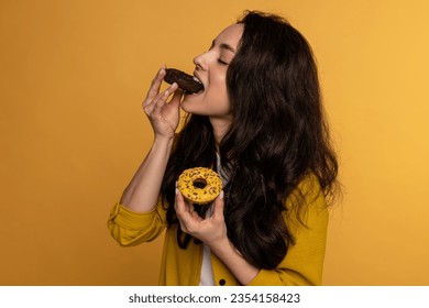 Portrait of a dark-haired woman biting off a piece of a chocolate-glazed biscuit while holding another in her hand. Self-indulgence and unhealthy food concept - Powered by Shutterstock