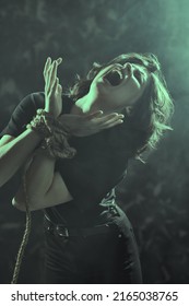 Portrait Of A Dark-haired Woman With Her Hands Tied Up With A Rope Suffering And Screaming In Severe Pain. Human Suffering.