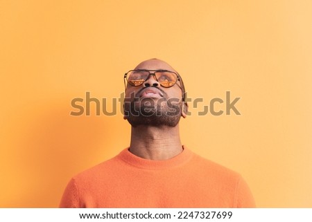 A portrait of a dark-skinned young man looking up in orange sunglasses, the man is wearing a sweater in the same color as the background. Orange color predominates, copyspace.