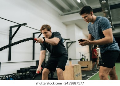 Portrait of dedicated sportsman exercising with battle rope while coach encouraging during cross training in health club standing with stopwatch. Concept of healthy lifestyle, physical sport activity. - Powered by Shutterstock