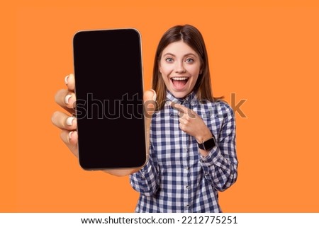 Portrait of delighted amazed young woman wearing checkered shirt pointing at smart phone with empty display, copy space for advertisement. Indoor studio shot isolated on orange background.