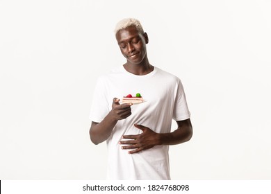 Portrait Of Delighted And Happy African-american Blond Guy Foodie, Rubbing Tummy And Smiling At Delicious Cake, Standing White Background