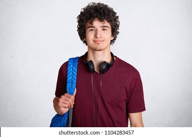 Portrait of delighted hipster male student with crisp hair, wears casual t shirt, carries backpack, has headphones on neck, ready to go for classes at university, isolated over white background