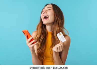 Portrait of delighted joyous caucasian woman holding cellphone and credit card isolated over blue background in studio