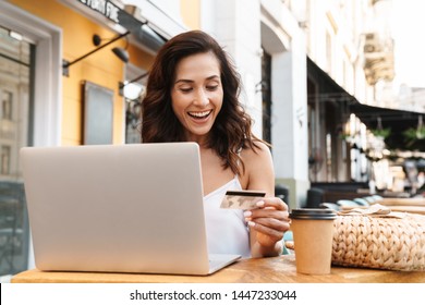 Porträt einer erfreuen verführerischen Frau mit Strohtasche, die eine Kreditkarte hält und einen Laptop benutzt, während sie im gemütlichen Café im Freien sitzt