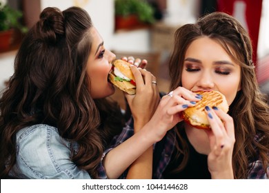Portrait of delighted teen girls with hairstyles enjoying delicious burgers in cafe.