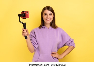 Portrait Of Delighted Woman Using Cell Phone And Steadicam For Broadcasting Livestream, Keeping Hand On Hips, Wearing Purple Hoodie. Indoor Studio Shot Isolated On Yellow Background.