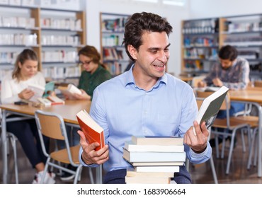 Portrait of delighted young man with stack of books in hands in public library