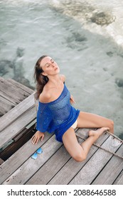 Portrait of a delightful deified young female model sitting and posing on a wooden bridge with her head tilted back, eyes closed and enjoying the atmosphere against the clear water. Privacy concept