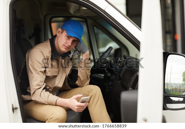 Portrait of delivery\
person in uniform holding mobile phone and looking at camera while\
sitting in the van