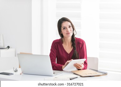 Portrait Of A Designer Working Home On New Ideas A Dark Hair Braided Woman Is Sitting At A White Table In Casual Clothes, She Is Looking At Camera, Holding Her Tablet And With Her Laptop Next To Her