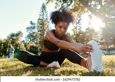 Portrait Of An Determined African American Young Woman Sitting On Green Grass In Morning Sunlight Stretching Her Legs Looking At Camera 