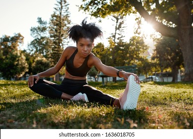 Portrait Of A Determined Fitness Young African American Woman Sitting On Green Grass Doing Exercise In The Park - Young Black Woman Warming Up Her Muscles Before Exercising 