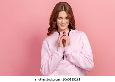 Portrait of devious cunning young woman with curly hair clasping hands and smirking mysteriously, scheming cheats, evil prank. Indoor studio shot, isolated on pink background