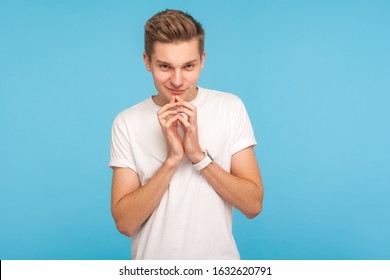 Portrait of devious man in casual white t-shirt holding hands together while thinking over clever cunning idea, smiling slyly and scheming prank. indoor studio shot isolated on blue background