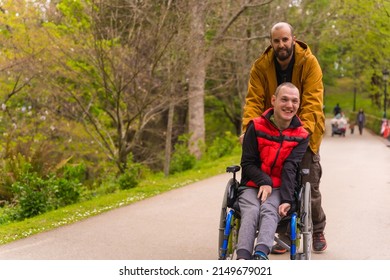 Portrait Of A Disabled Person In A Public City Park In The Wheelchair, Walking Along A Beautiful Park Path In Spring