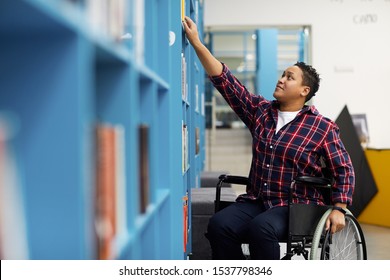 Portrait of disabled student in wheelchair choosing books while studying in college library, copy space