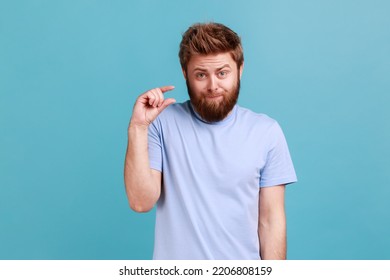 Portrait of disappointed bearded man showing a little bit gesture, dissatisfied with low rating, measuring scale, frowning face and looking at camera. Indoor studio shot isolated on blue background.