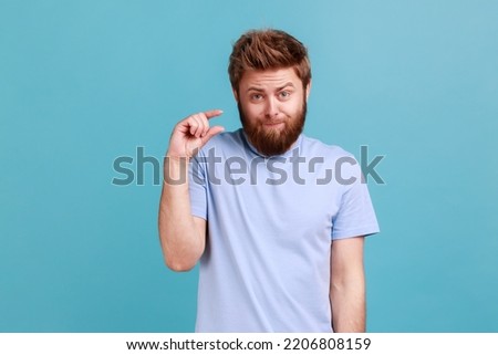 Portrait of disappointed bearded man showing a little bit gesture, dissatisfied with low rating, measuring scale, frowning face and looking at camera. Indoor studio shot isolated on blue background.