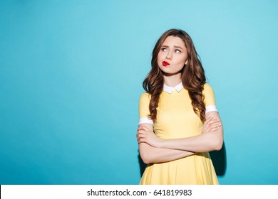 Portrait of a disappointed cute girl in dress standing with arms folded and looking away isolated over blue background
