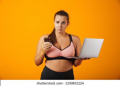 Portrait Of A Disappointed Overweight Young Woman Wearing Sport Clothing Standing Isolated Over Yellow Background, Using Laptop Computer, Holding Chocolate Bar