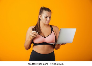 Portrait Of A Disappointed Overweight Young Woman Wearing Sport Clothing Standing Isolated Over Yellow Background, Using Laptop Computer, Holding Chocolate Bar