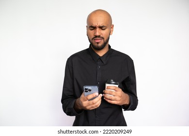 Portrait Of Disappointed Young Man Using Mobile Phone Against White Background. Bearded Businessman Wearing Black Shirt Holding Coffee Cup And Reading Bad News On Smartphone. Bad News Concept