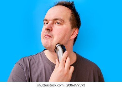 Portrait Of Disheveled Adult Man Shaving Stubble With Electric Razor, Blue Studio Background