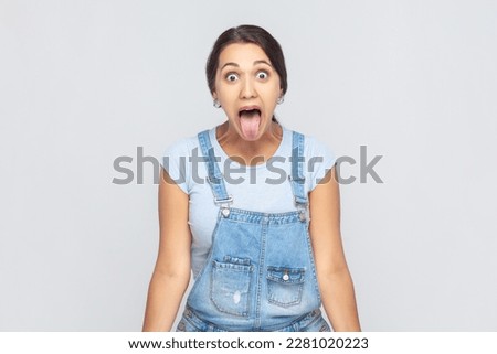 Portrait of disobedient woman wearing denim overalls sticking out tongue and grimacing, aping showing derisive, naughty expression. Indoor studio shot isolated on gray background.