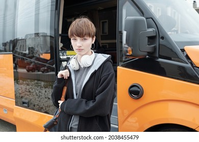 Portrait Of Displeased High School Caucasian Girl With Wireless Headphones Around Neck Standing Against Open Bus Door