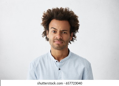 Portrait of displeased mixed race attractive man with dark narrow eyes and curly thick hair wearing formal shirt frowning his face raising up eyebrows with discontent having some problems at his work