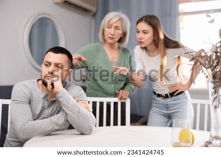 Portrait of displeased young adult man sitting at home ignoring his wife and mother-in -law berating him
