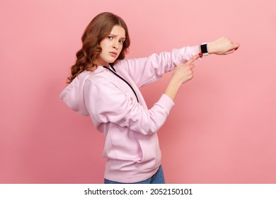 Portrait of displeased young brunette female in hoodie looking impatient and pointing wristwatch, warning of deadline, late hour. Indoor studio shot isolated on pink background - Shutterstock ID 2052150101