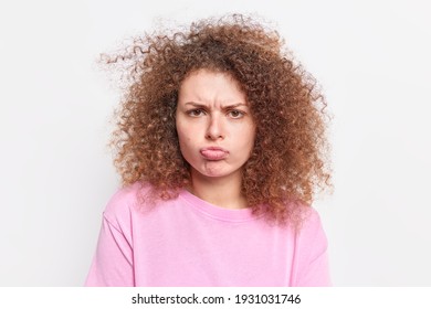 Portrait of dissatisfied upset curly haired woman looks with sullen expression grimaces from hearing offensice words looks with dislike at camera wears casual pink jumper isolated on white background