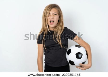 Portrait of dissatisfied young caucasina girl holding a football ball over white background smirks face, purses lips and looks with annoyance at camera, discontent hearing something unpleasant