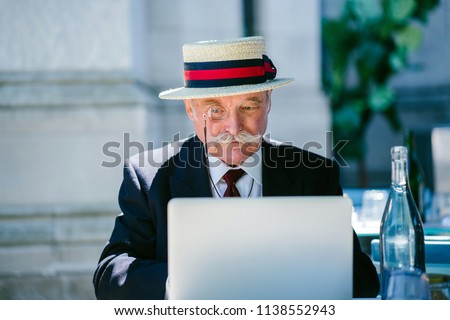 Portrait of a distinguished elderly Caucasian (European, British) gentleman working on his laptop computer during the day at a cafe. He is wearing a suit and tie, monocle and hat and has a moustache.