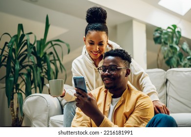 Portrait of a diverse couple relaxing on cozy sofa at home using smartphone and drinking hot beverages. African american man and hispanic woman sitting on couch while chatting and talking. Copy space - Powered by Shutterstock