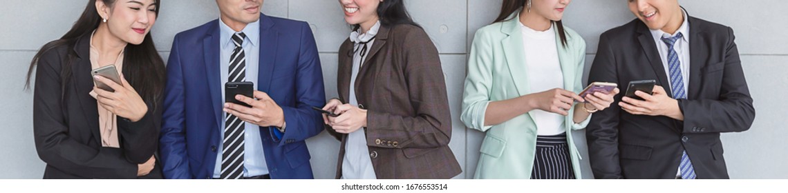 Portrait Of Diverse Culture Asian And Caucasian Business People Watching Smart Mobile Phones Against Office Gray Wall. Young People Addiction To New Technology. Community Business Communication Banner